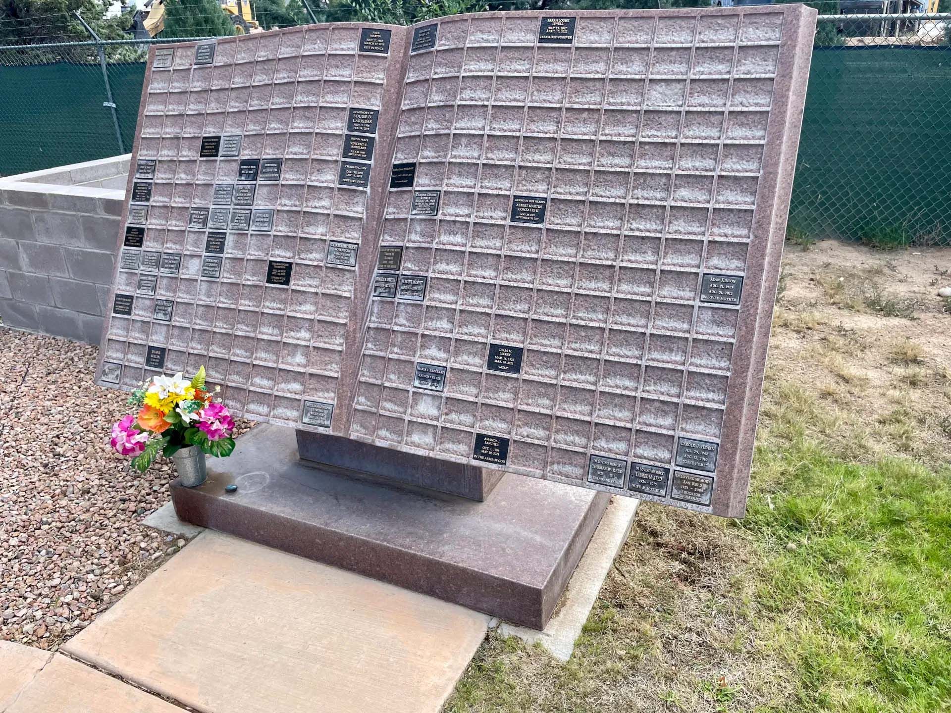 Memorial wall with small plaques, a granite base, and flowers in an outdoor setting.
