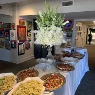 Buffet table with pasta, pizzas, and a tall white floral arrangement in a gallery-like room