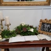 Decorated wooden table with greenery, candles, open book, and a small chalkboard sign reading “sign in”