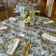Round banquet table with blue-and-white floral tablecloth, folded napkins, gold cutlery, and a floral centerpiece.