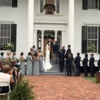 Wedding party gathered on the front steps of a white house, with bride and groom at center.