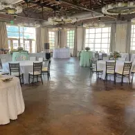 Empty event hall with round tables, white chairs, and large windows under exposed industrial ceiling