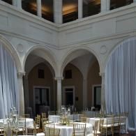 Elegant courtyard dining setup with round tables and white chairs under arched colonnades and draped curtains