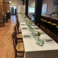 Long banquet table set with white linens, plates, and greenery in a restaurant dining room