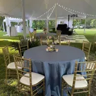Round table with gold chairs under a white tent at an outdoor event lawn