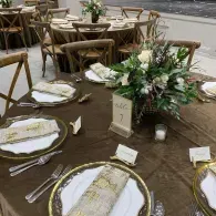 Elegant outdoor table setting with gold-rimmed plates, wicker chairs, and a floral centerpiece.