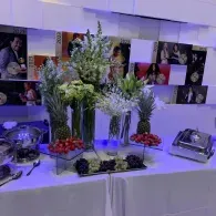 Buffet table with floral centerpieces, fruit, and silver chafing dishes under purple lighting