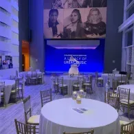 Blue-lit banquet hall with round tables and gold chairs, stage backdrop reading “City of Dreams Music”