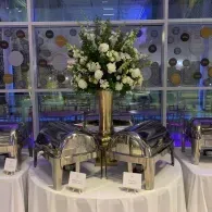 White floral arrangement on a banquet table with silver chafing dishes at an event venue.