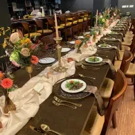 Elegant long banquet table set with dark plates, gold accents, flowers, and candles in a restaurant