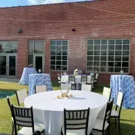Outdoor patio with round white table, chairs, and blue checkered tablecloths in front of a brick building