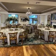 Elegant banquet room with round, white-clothed tables, gold chairs, chandelier, and hardwood floor.