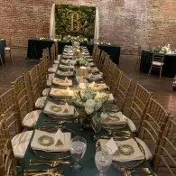 Long banquet table with gold chairs, black runner, floral centerpieces, and brick wall backdrop