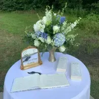 Outdoor memorial table with white cloth, flower arrangement, guest book, and framed sign