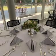 Round banquet table set with folded napkins, glassware, silverware, and a floral centerpiece by large windows