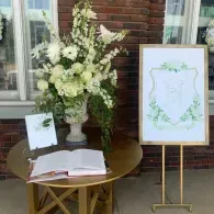White floral arrangement, guestbook, and framed sign on a gold table beside a brick wall.