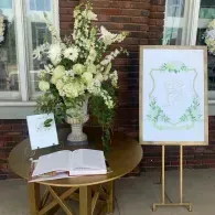 Elegant outdoor memorial table with white flowers, an open book, and framed sign beside a brick wall
