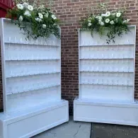 Two white display shelves with floral arrangements on top, set against a brick wall outdoors