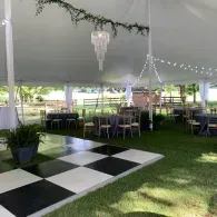 Outdoor event tent with black-and-white checkered dance floor, white draping, and banquet tables on grass.