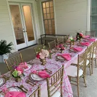 Decorated outdoor dining table with pink floral tablecloths and gold chairs on a covered patio