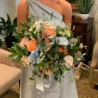 Person holding a white, peach, and green bouquet with ribbon, wearing a pale blue dress indoors