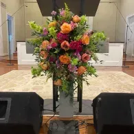 Colorful floral arrangement on a stand at a church or auditorium stage, framed by speakers and a podium.