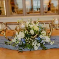 Rustic table centerpiece with white and blue flowers, candles, and antlers on a wooden table