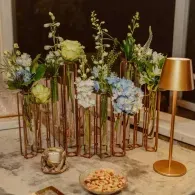 Elegant floral arrangement with blue and green flowers in tall copper vases beside a gold lamp and snacks on a table