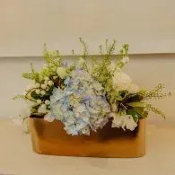 Floral arrangement with pale blue, white, and green flowers in a rectangular copper vase on a wall shelf