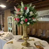 Tall brass vase with red and white flowers on a white-clothed dining table in a rustic room