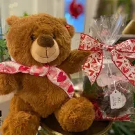 Brown teddy bear with a pink scarf beside a red-and-white wrapped gift basket