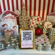 Festive holiday table display with QR code sign, striped backdrop, ornaments, candles, and wrapped gifts