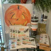 Halloween display with orange jack-o'-lantern decor, “Broom Shop” items, and striped mugs on a table