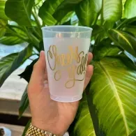 Hand holding a frosted cup with gold script in front of large green leaves