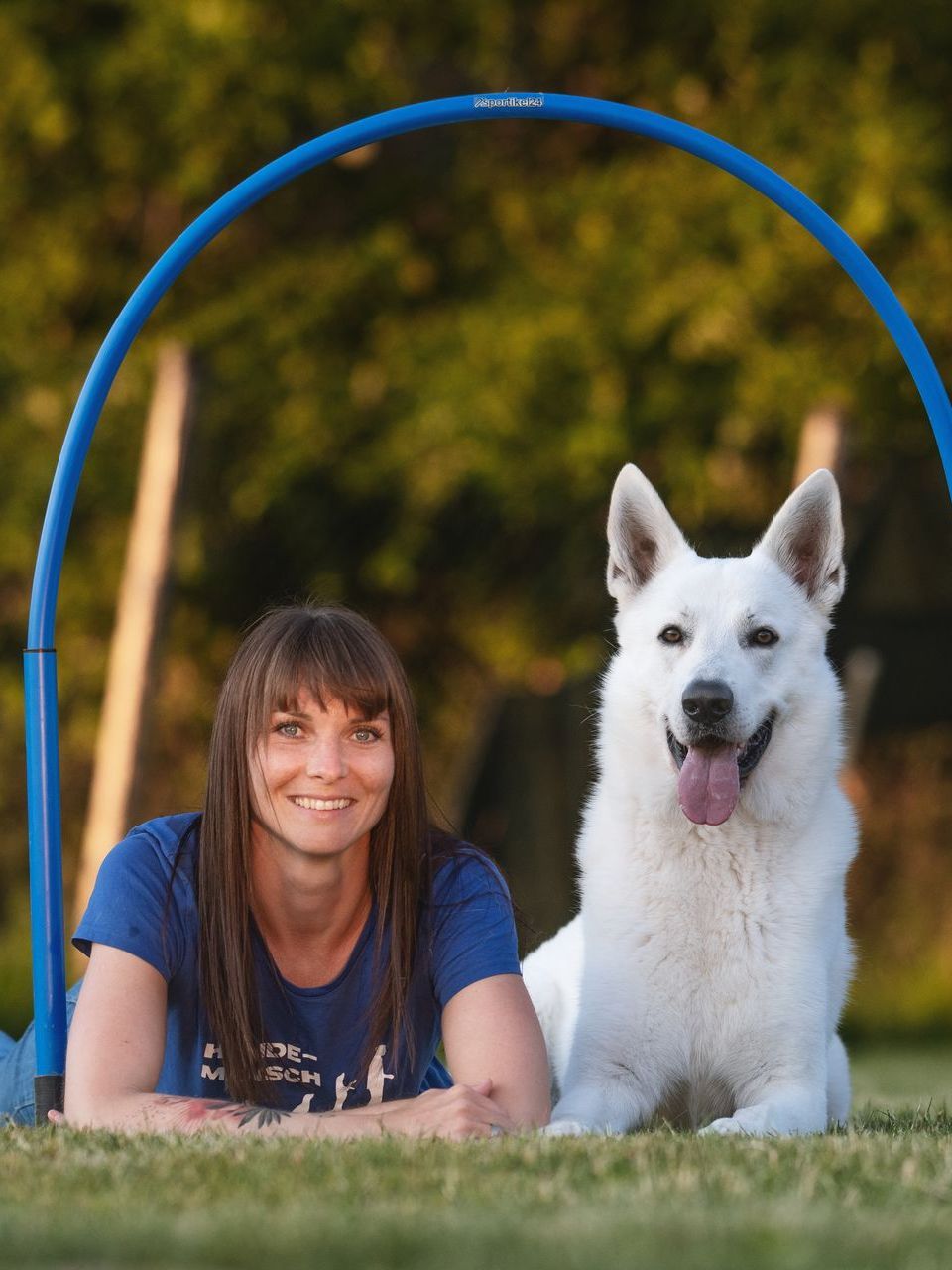 Ramona Houscht und weißer Schäferhund Jimmy liegen auf der Wiese unter einem Hoop und blicken fröhlich in die Kamera