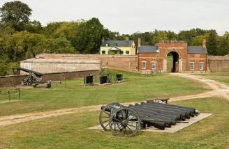 Historic fort with brick gate, grassy field, cannon, and old buildings under trees Historic fort with brick gate, grassy field, cannon, and old buildings under trees