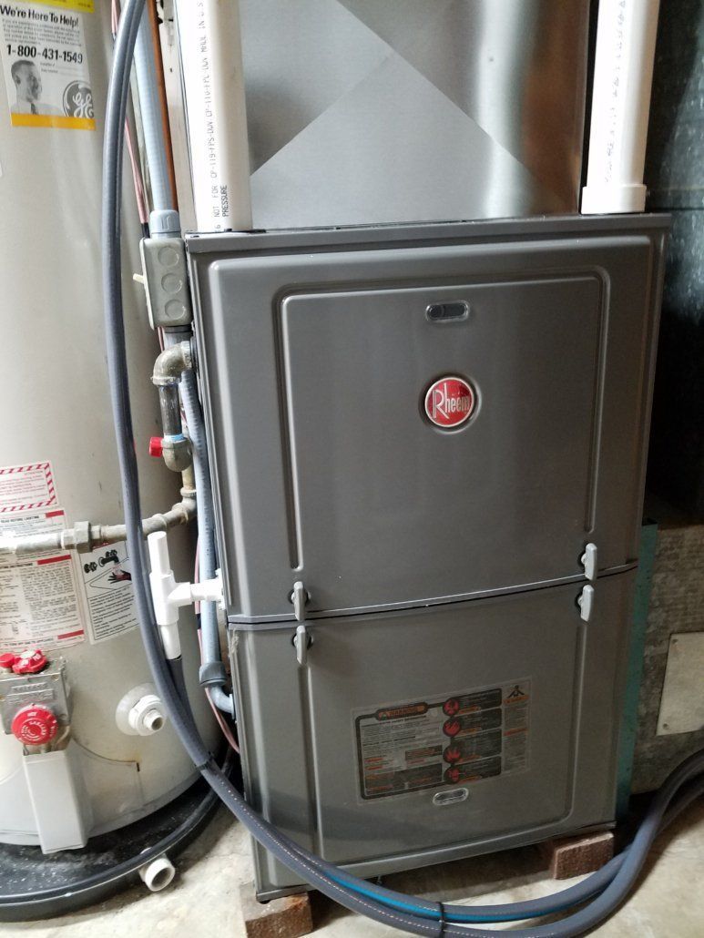 Gray furnace and water heater in a utility closet with exposed pipes and hoses.