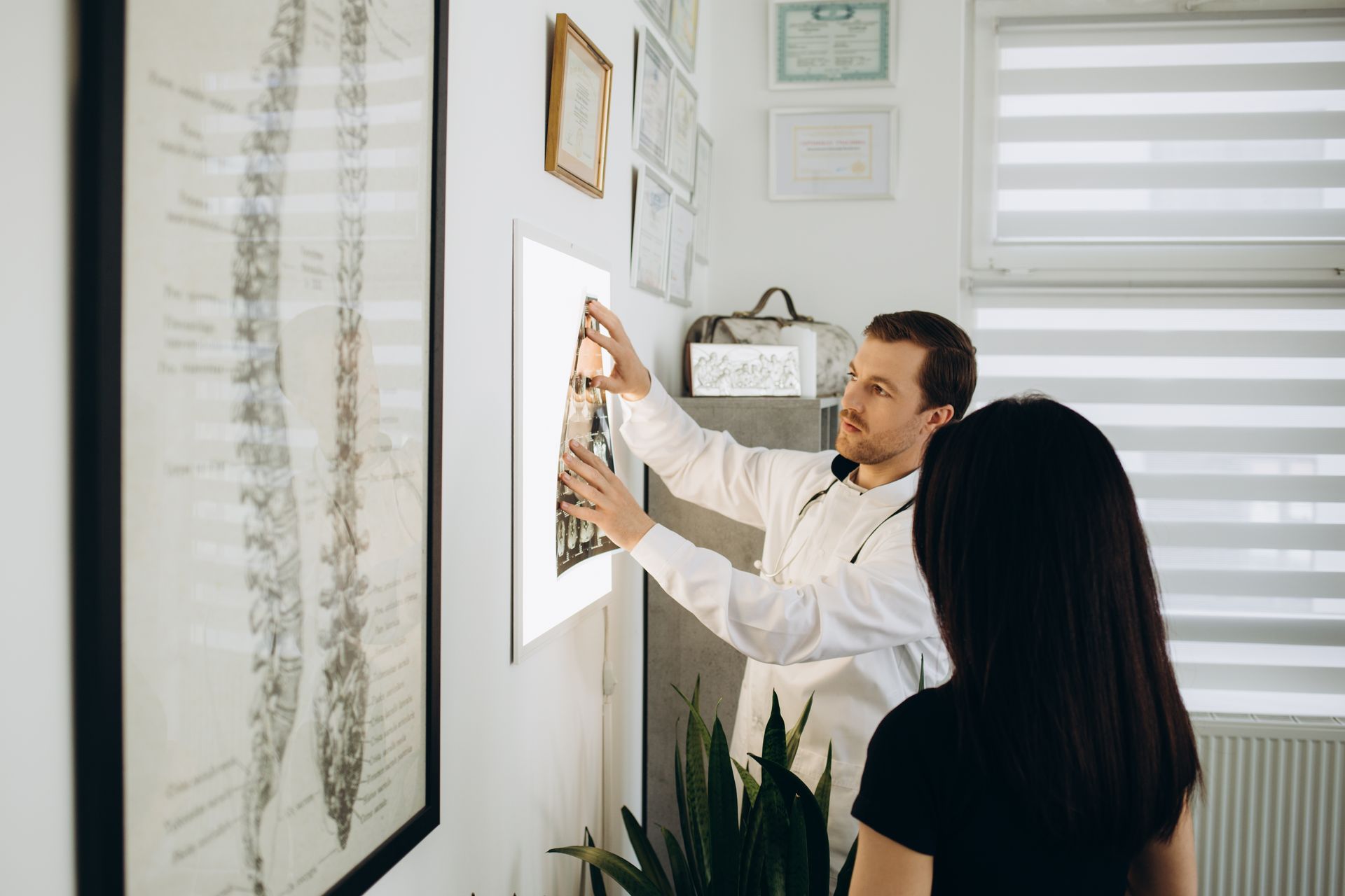 A chiropractor pointing at X-ray, explaining to patient. Office with spinal chart on the wall.