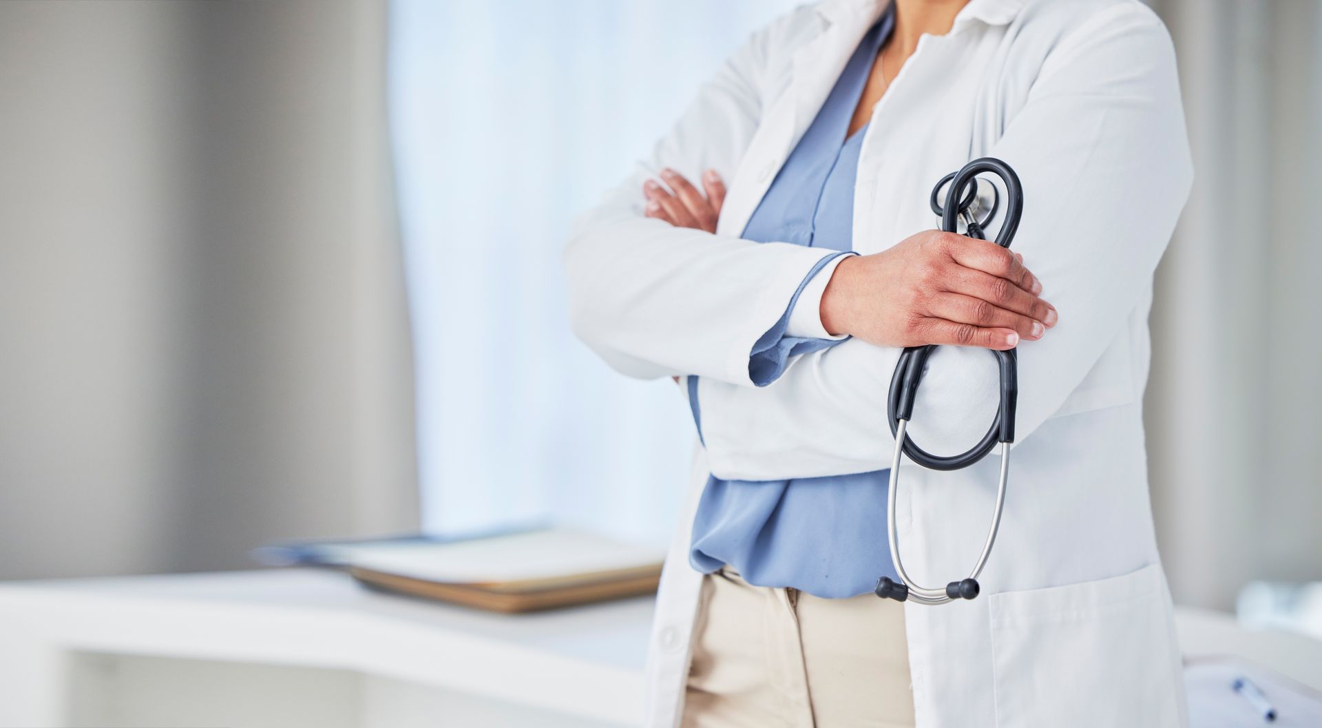 Person in white lab coat holding stethoscope, arms crossed.