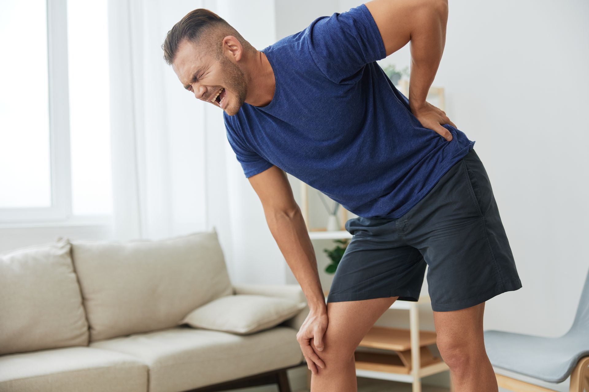 Man in blue shirt and shorts clutches his lower back and knee in pain indoors.