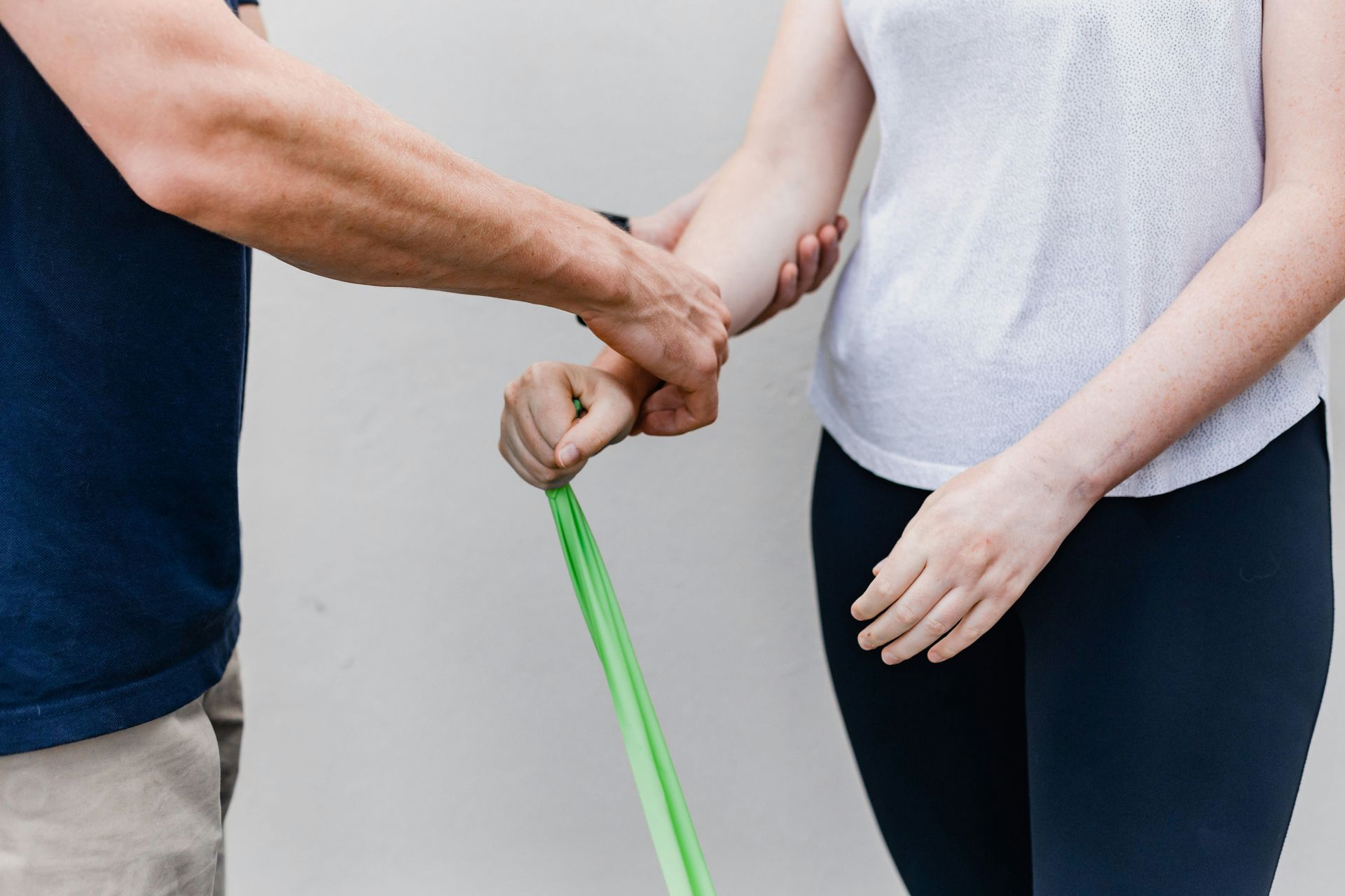 Person assisting another with arm exercise using a green resistance band against a white wall.