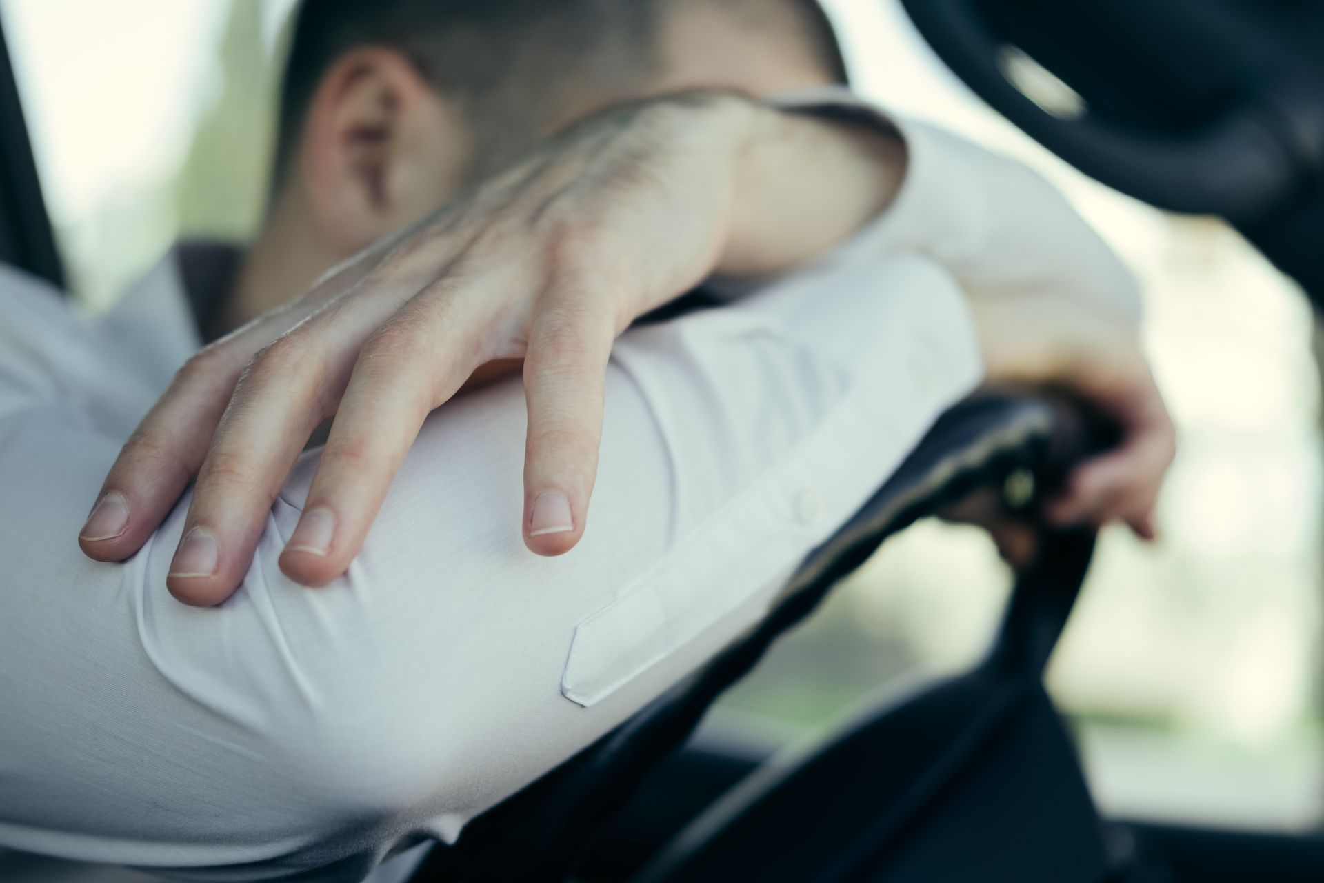 Man resting his head on arms, leaning on steering wheel in a car, looking exhausted.