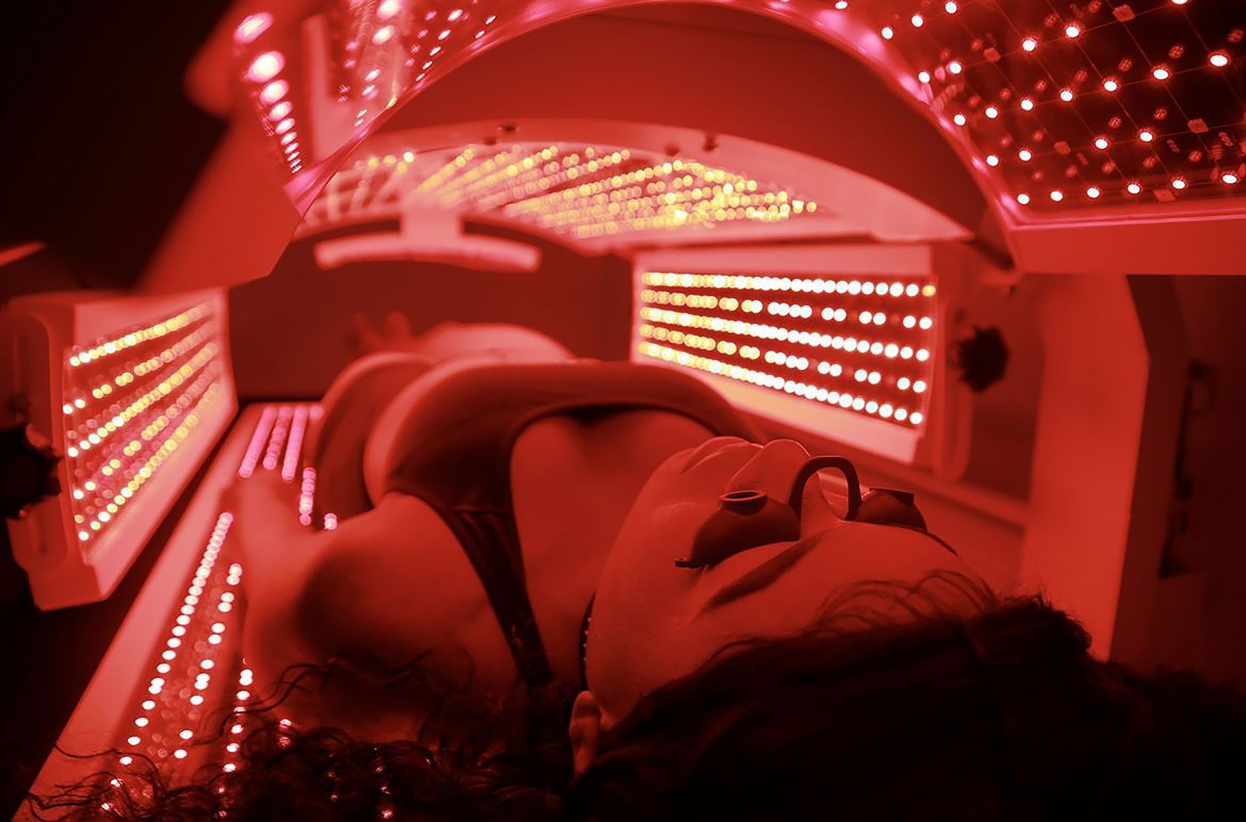 Person lying inside a red-lit tanning or infrared therapy booth, with glowing panels around them