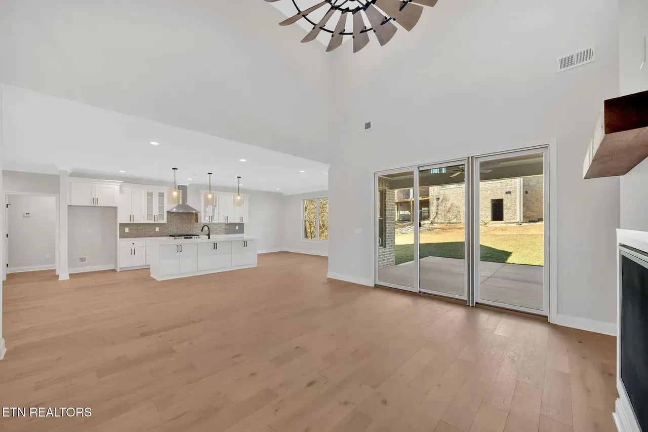 Open-concept living space with kitchen and sliding glass doors to a patio. Light wood floors, white walls, and a windmill ceiling fan.