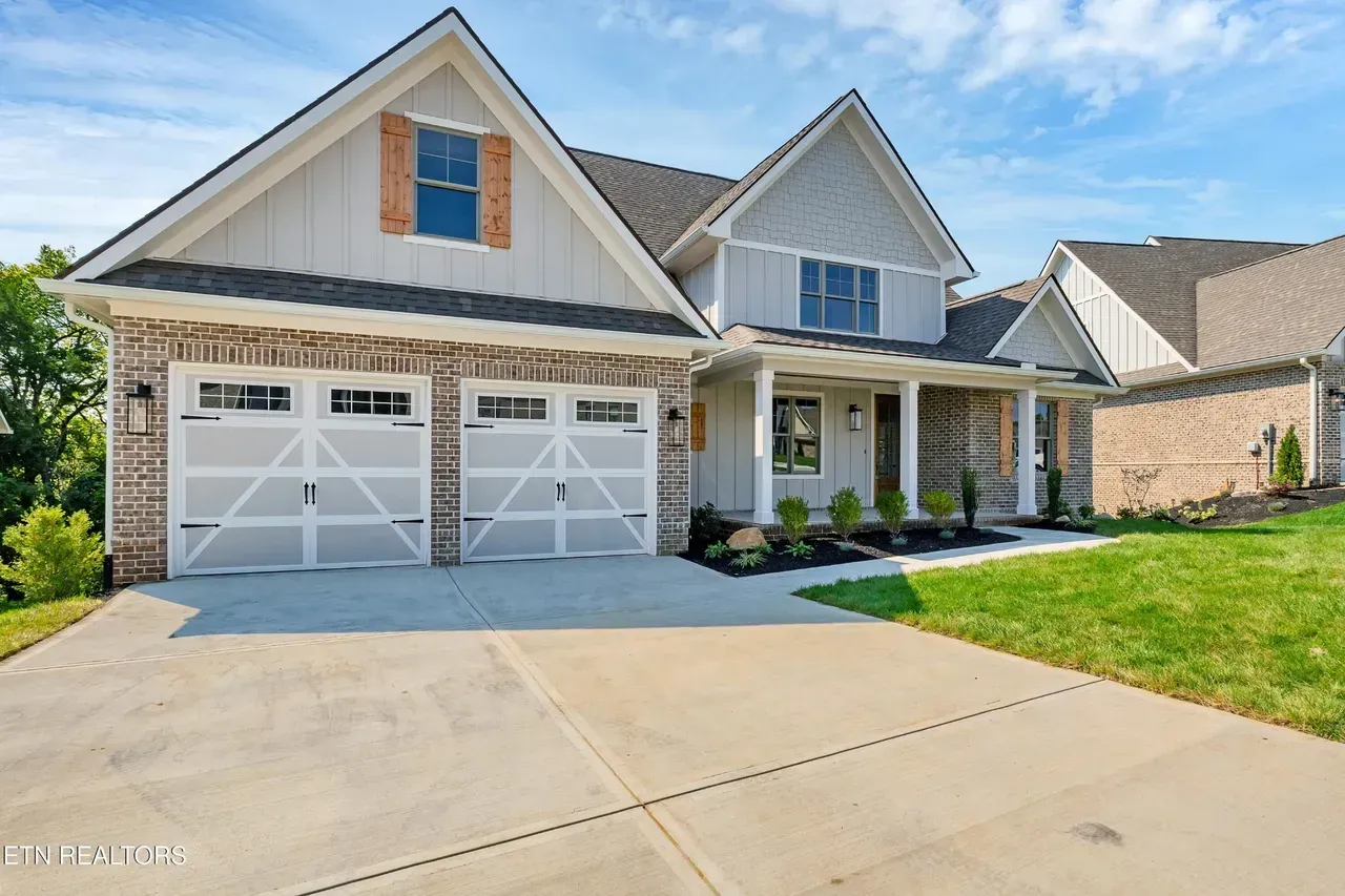 Exterior view of a two-story house with a brick facade, white garage doors, and a concrete driveway on a sunny day.