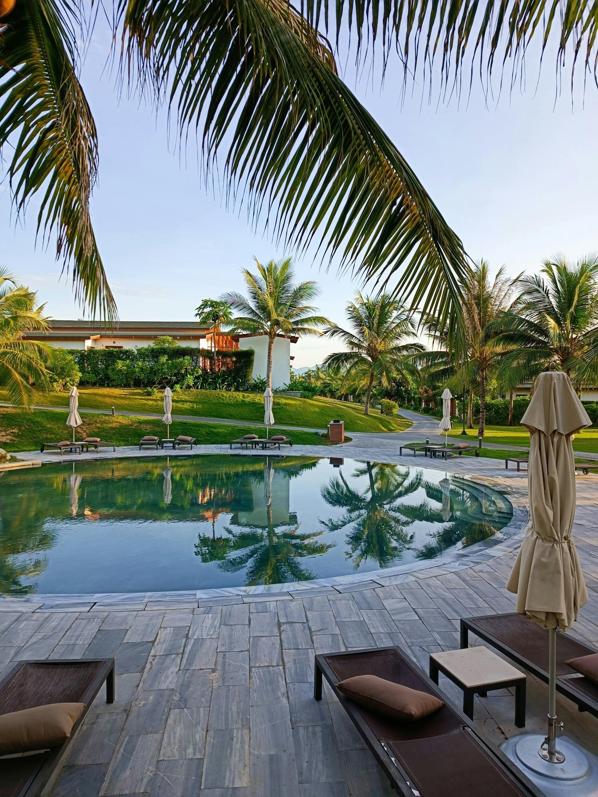 Piscina de resort tropical com palmeiras, espreguiçadeiras e um guarda-sol fechado com vista para as águas calmas.