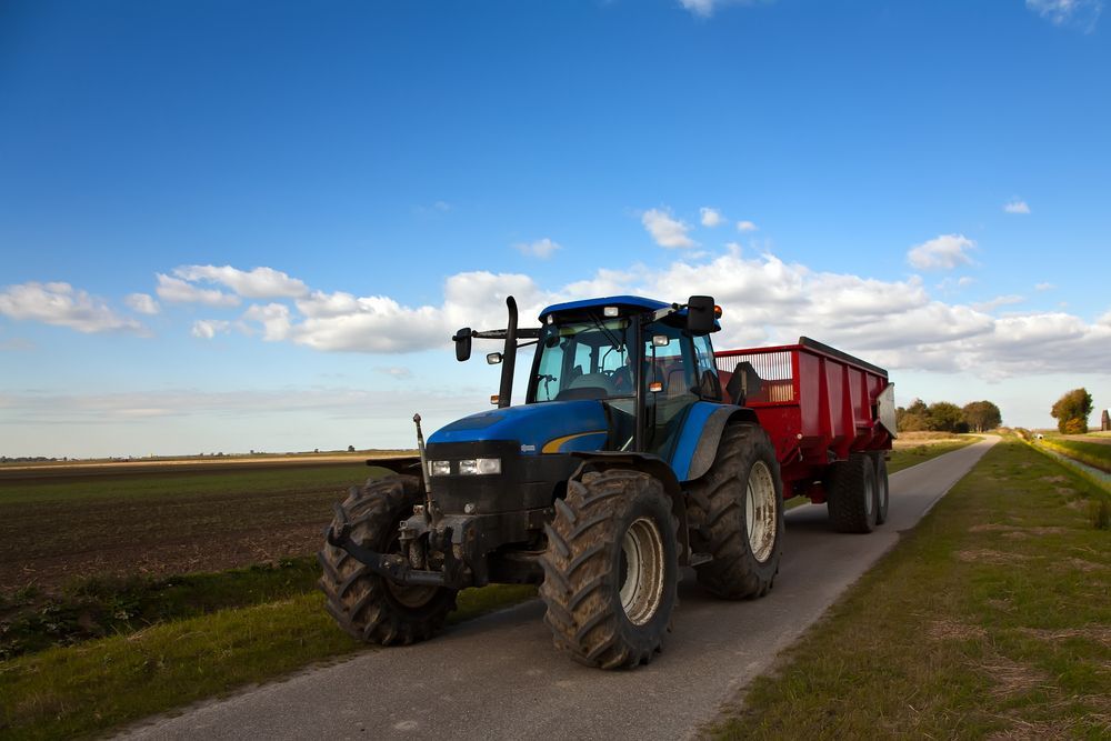 A blue tractor is pulling a red trailer down a road. — Nabiac Mower & Tractor Repairs in Nabiac, NSW