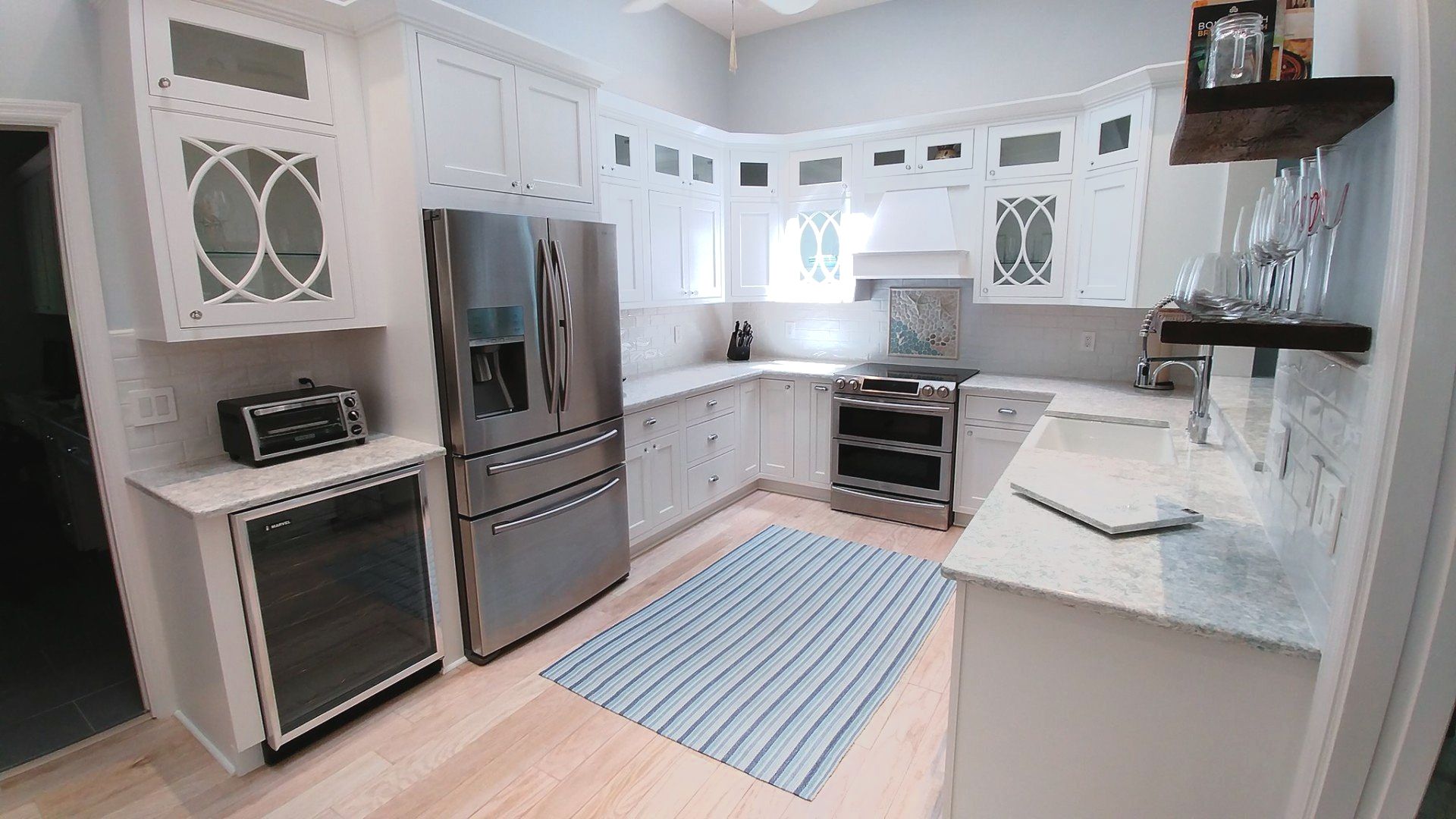 White kitchen with stainless steel appliances, blue striped rug, and wood shelving.