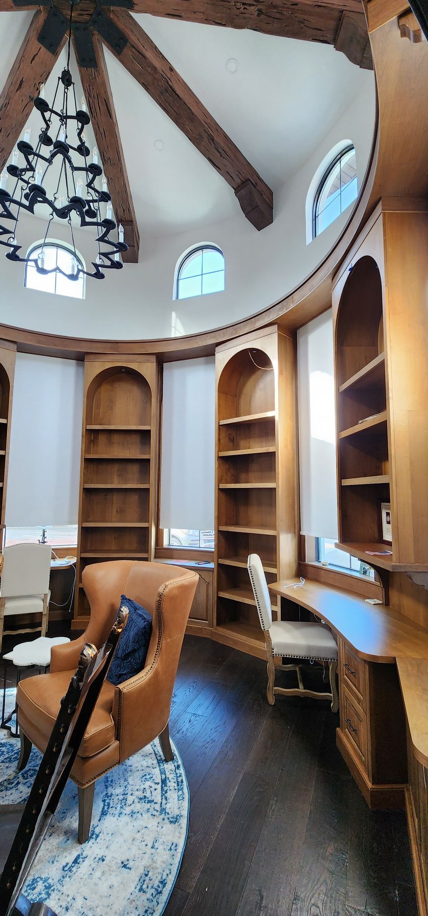 A circular library with wooden shelves, desk, and an armchair. Dark wood floors, a patterned rug, and a chandelier are visible.