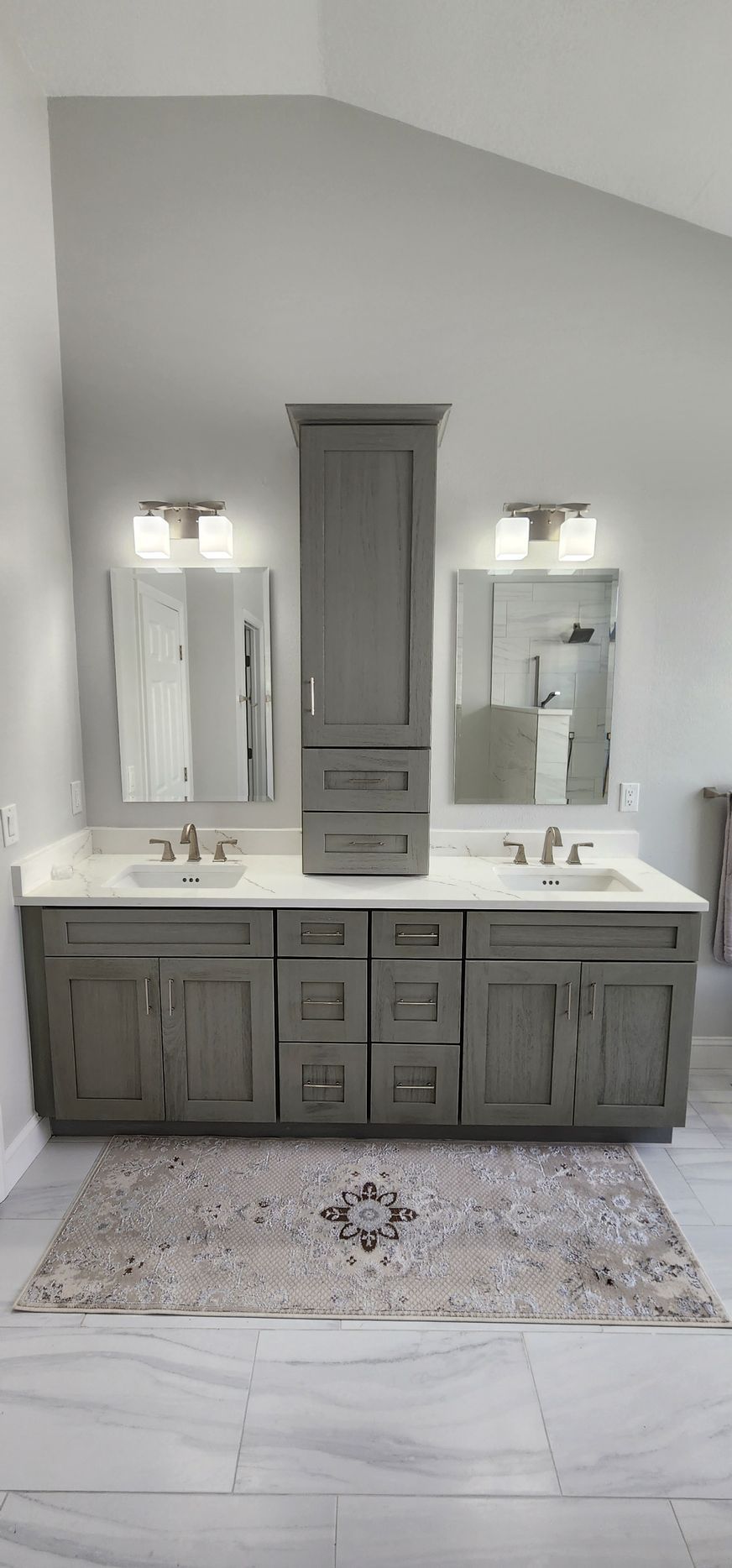 Gray bathroom vanity with mirrors, lights, and a rug. A tall cabinet is centered.